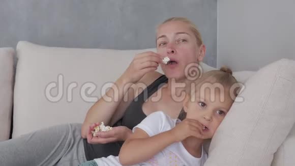 一个女人和一个小女孩坐在柔软的沙发上吃着爆米花看着电视屏幕视频的预览图