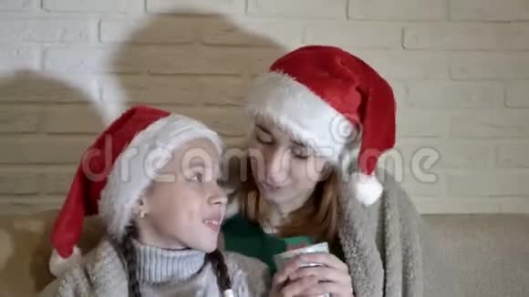 两个戴着圣诞老人帽子的女孩用毯子盖着的肖像她们坐在沙发上拿着杯子微笑着关闭视频的预览图