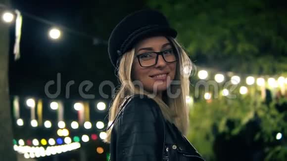 一位戴着时尚帽子时尚眼镜和一件黑色皮夹克的漂亮女孩在夜间公园散步的肖像光线视频的预览图