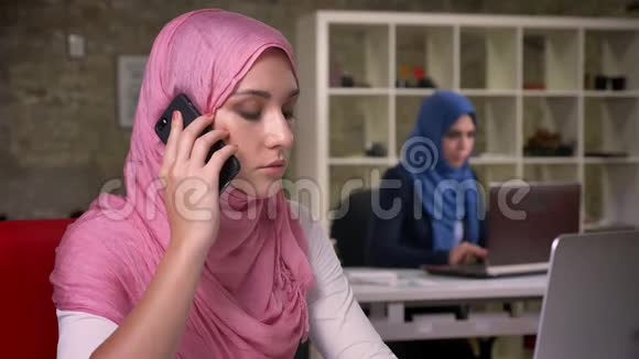 自信的好穆斯林女孩专注于在她的脑海中电话交谈坐在砖办公室与其他阿拉伯人视频的预览图