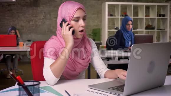 穿着粉色头巾的漂亮阿拉伯女人在办公室里自信地坐着一边打电话一边冷静和放松视频的预览图