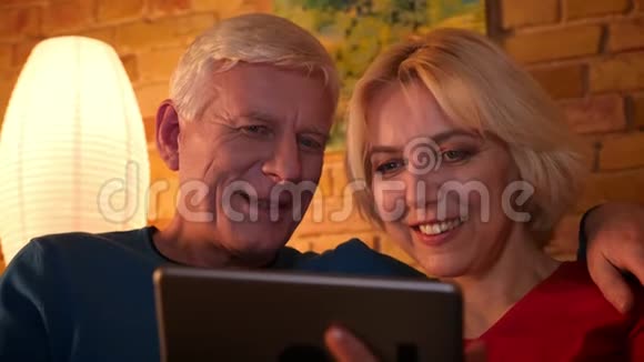 在舒适的室内沙发上坐在一起休息的老夫妇在平板电脑上浏览网页的特写镜头视频的预览图