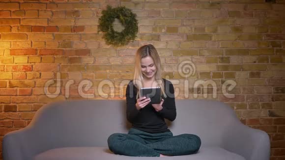使用平板电脑坐在舒适的家里的沙发上的年轻漂亮金发女性的特写照片视频的预览图