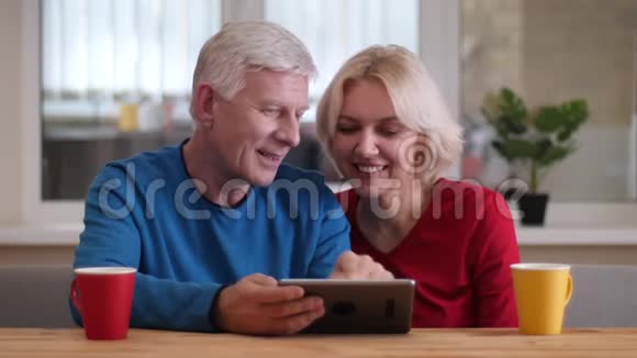 在舒适的公寓里一对幸福的老夫妇在桌上放着茶杯的平板电脑上近距离拍摄视频的预览图
