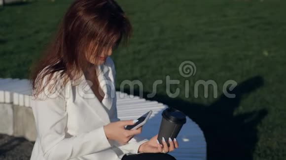有智能手机的年轻女人坐在公园里带电话的白色休闲套房漂亮女孩的时尚商务照片视频的预览图