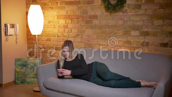 一位年轻漂亮的金发女性使用电话躺在舒适的室内沙发上的特写照片视频的预览图