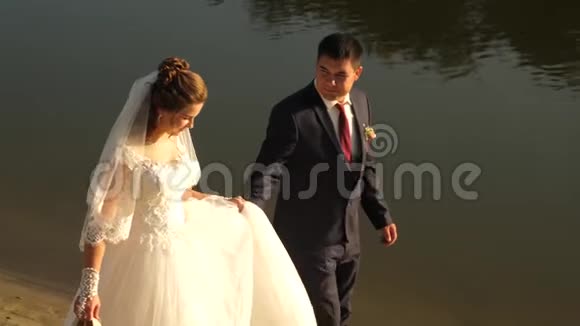 幸福的夫妇沿着河岸散步可爱的新郎穿着西装新娘穿着白色连衣裙在海滩上散步一种概念视频的预览图