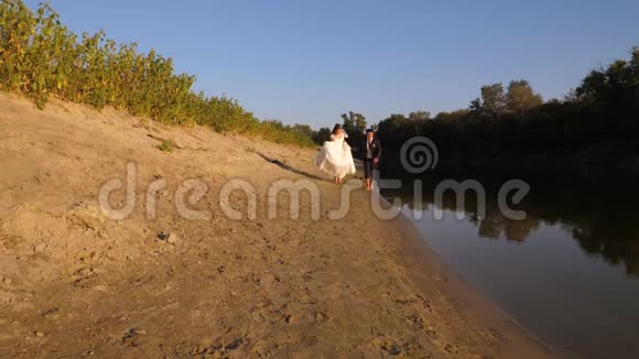 一对恋爱中的情侣沿着河边跑快乐的新郎穿着西装新娘穿着白色连衣裙在海滩上奔跑幸福的概念视频的预览图
