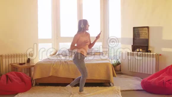 快乐的黑发年轻女子戴着耳机独自在卧室里跳舞大窗户在智能手机上听音乐女孩视频的预览图