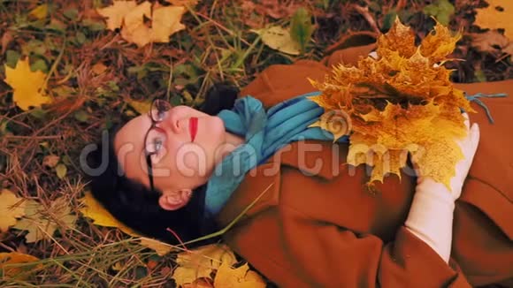 一个戴眼镜的年轻女子躺在一个秋天公园的草地上枫叶正落在她的上面视频的预览图