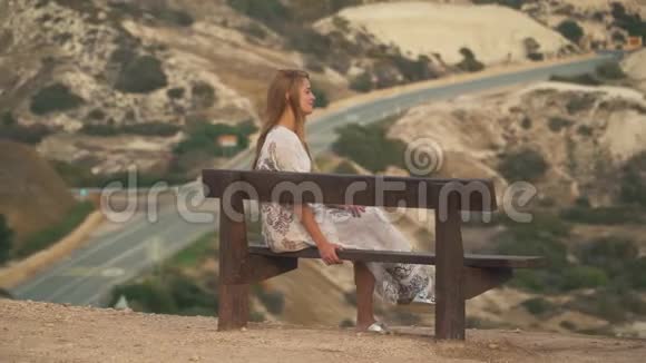 孤独的女孩穿着白色的长裙坐在长凳上美丽的岩石山丘和道路在背景中休闲视频的预览图