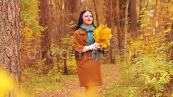 秋天公园散步时一个穿着外套的年轻女子欣赏手中的枫叶慢动作视频的预览图