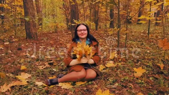 戴眼镜的年轻快乐女子手里拿着枫叶坐在秋天公园的草地上视频的预览图