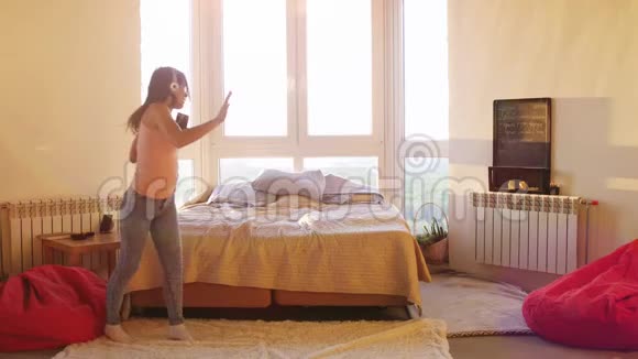 幸福的年轻美女戴着耳机独自在房间里跳舞窗户很大听着智能手机上的音乐女孩摔倒了视频的预览图
