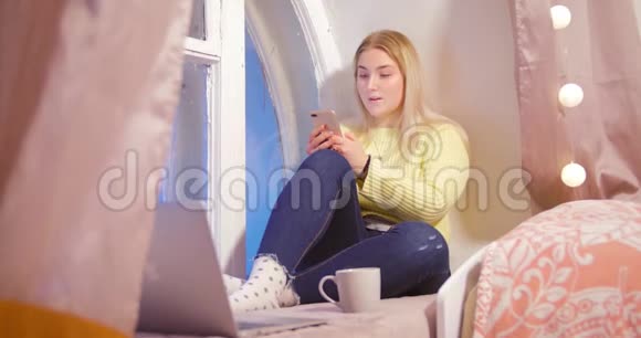 一个穿黄色毛衣的金发女孩正坐在房间窗户附近的电话旁背景是粉红色视频的预览图