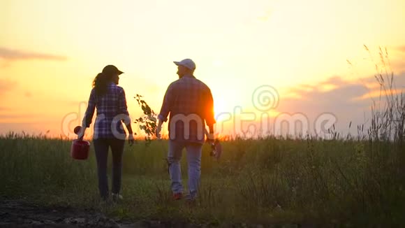 农民男女农民夫妇在日落或日出时在田野散步搬运树苗浇水罐和铲子视频的预览图