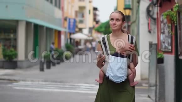 妈妈带着婴儿带着吊带走在街上去西班牙旅游并在她的手机上拍照视频的预览图