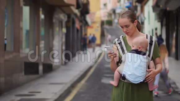 妈妈站在街上一个婴儿穿着吊带看着手机屏幕看着照片视频的预览图