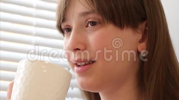年轻的女孩喝着白色玻璃早咖啡或茶站在窗边微笑好心情视频的预览图