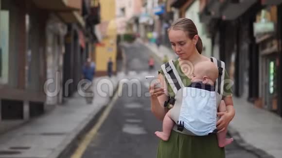 妈妈站在街上带着一个穿着吊带的孩子看着手机屏幕在社交中交流视频的预览图