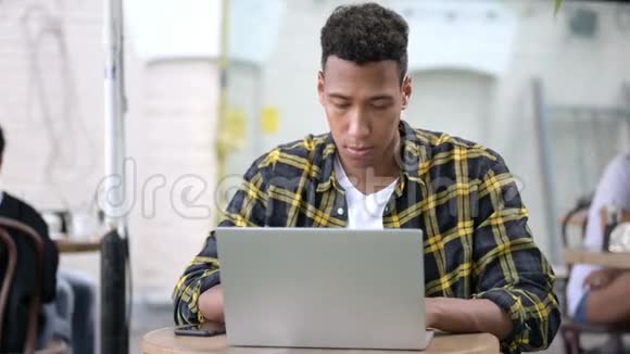 带头痛的非洲青年男子使用笔记本电脑户外咖啡厅视频的预览图