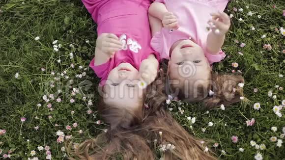 两个可爱的小女孩倒躺在草地上和鲜花上做着空中亲吻和挥手视频的预览图