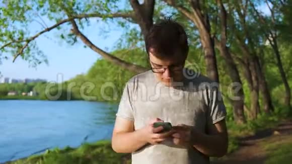 年轻人沉迷于他的手机在森林里沿着河走看着他的设备库存录像男子视频的预览图