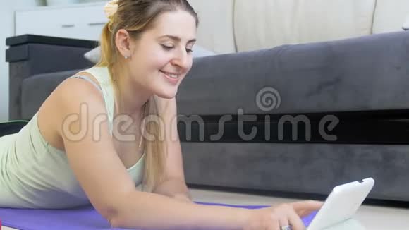 美丽微笑的年轻女子躺在健身垫上使用数字平板电脑的4k视频女生看健身课视频的预览图