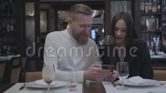 坐在餐桌旁的餐厅里的金发男人和漂亮的黑发女人那个男人给他女朋友看视频的预览图