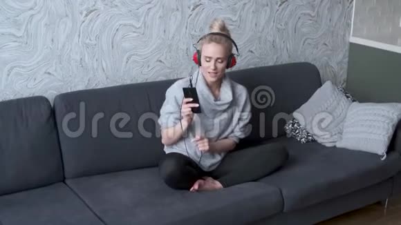 可爱的中年金发女人坐在家里的沙发上听音乐视频的预览图