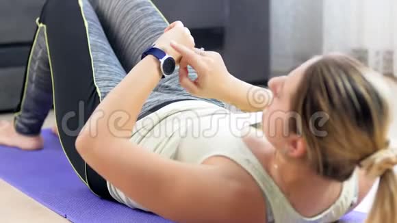 视频中年轻女子躺在瑜伽垫上在做腹肌紧缩前在手腕上安装健身跟踪器视频的预览图