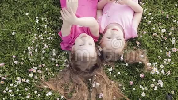 两个可爱的小女孩倒躺在草地上和鲜花上做着空中亲吻和挥手视频的预览图