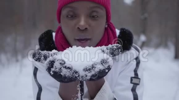 非洲裔美国女孩穿着温暖的衣服戴着豆帽和白色夹克把雪花从手中吹入画中视频的预览图