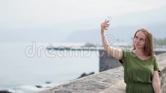 一个穿着绿色衣服的漂亮女孩站在海边的手机上自拍微笑着视频的预览图