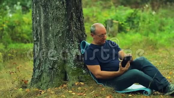 戴眼镜的人坐在公园的一棵树下在平板电脑上工作视频的预览图