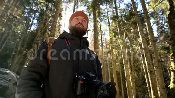 一位留着胡子的严肃摄影师的肖像他背着背包和一架照相机站在针叶林中视频的预览图