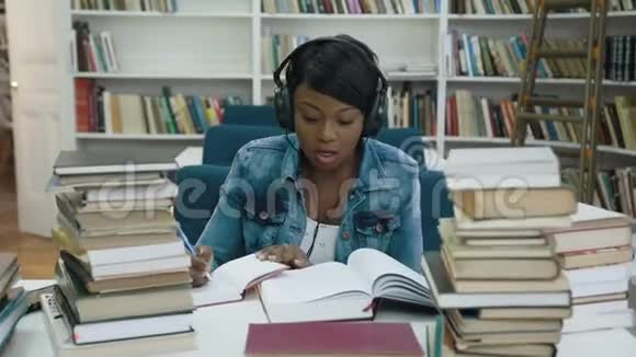有魅力的非洲年轻女性在耳机里听音乐同时从笔记本上为大学做一些笔记视频的预览图