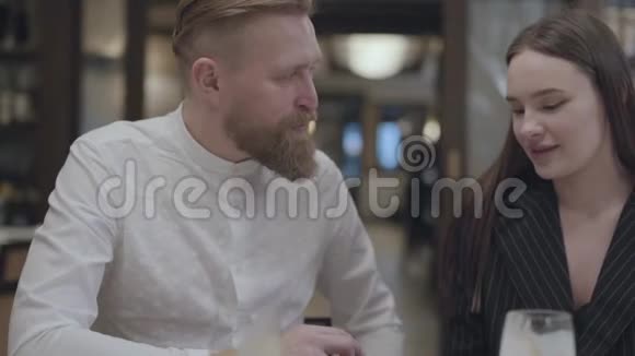 有胡子的男人和漂亮的黑发女人坐在餐桌旁的餐馆里他展示了他女朋友的信息视频的预览图