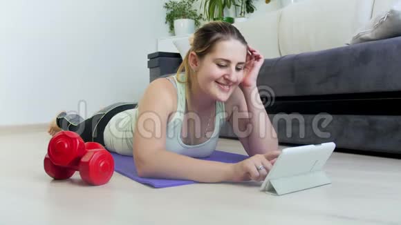 美丽微笑的年轻女子躺在健身垫上使用数字平板电脑的4k视频女生看健身课视频的预览图