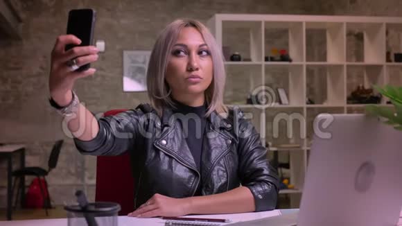 可爱热情的白种人金发女人坐在桌面上坐在室内一边用手机自拍视频的预览图