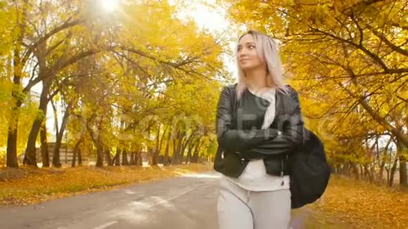 年轻漂亮的女孩走在秋天的跑道上一个穿着皮夹克和运动服的年轻时髦女人走在沥青路上视频的预览图
