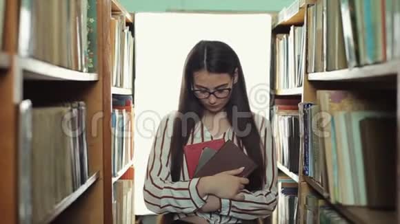 长着黑色长发的漂亮女孩直视镜头在图书馆的书架上漫步视频的预览图
