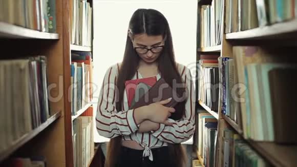 长着黑色长发的漂亮女孩直视镜头在图书馆的书架上漫步视频的预览图