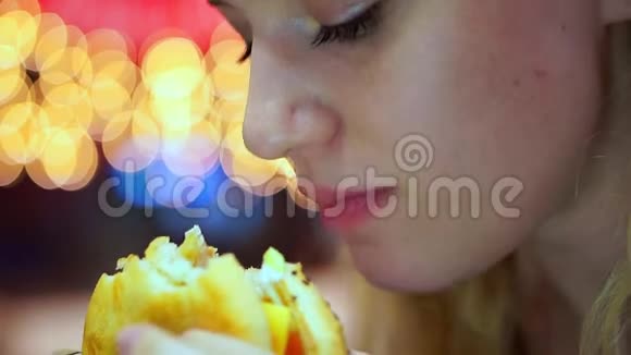 特写镜头年轻的少女在咖啡馆里吃一个多汁的汉堡包快餐晚上在灯泡的背景上视频的预览图