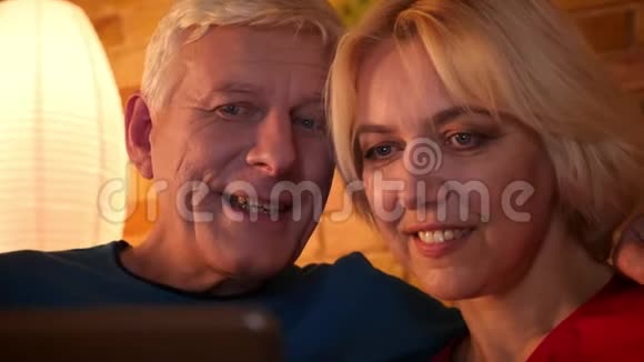 一对老年幸福夫妇在平板电脑上浏览网络的特写镜头他们坐在沙发上在室内休息视频的预览图