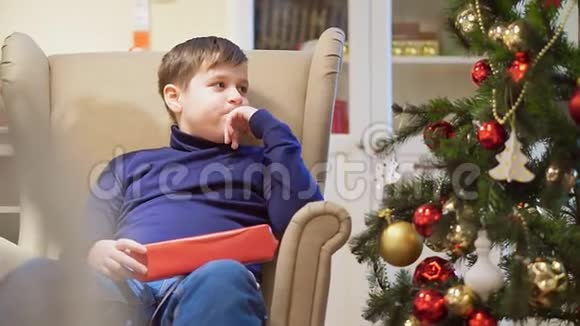 胖乎乎的小男孩坐在圣诞树附近的椅子上他拿着一个红色盒子里的礼物视频的预览图