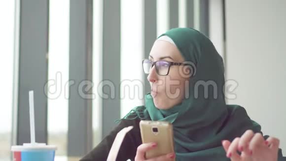 年轻的穆斯林妇女戴着头巾坐在靠窗的桌子旁用电话和土豆视频的预览图