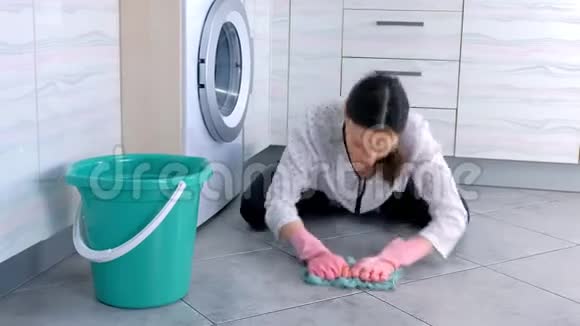 穿着粉红色橡胶手套的疲惫的女人用一块布擦着厨房的地板地板上的灰色瓷砖视频的预览图