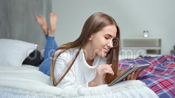 快乐美丽的女孩享受周末躺在床上使用平板电脑视频的预览图