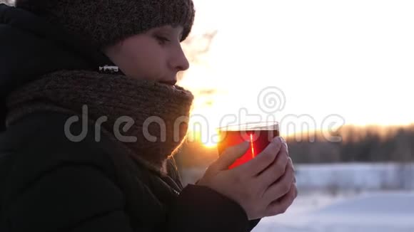 冬天的女孩穿着衣服手里拿着透明的玻璃在夕阳下的冬天公园里喝着热茶女孩温暖她的手视频的预览图